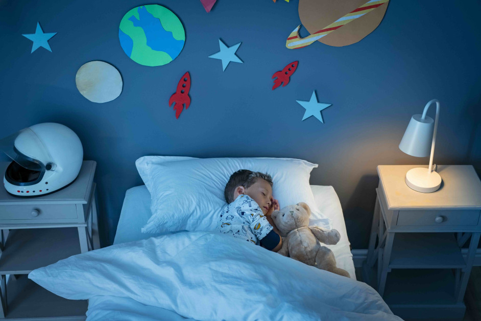 High angle view of little boy dreaming of becoming an astronaut while sleeping with teddy bear in space decorated room. Top view of dreamer child sleeping on bed with solar system and planet decoration during the night. Cute kid with astronaut helmet on side table and the light on.