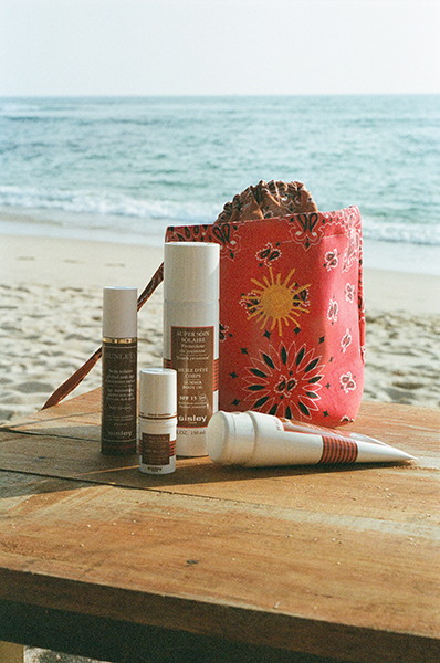 Photo du sac Sisley x Call It By Your Name et des produits solaire sur un table en bois avec la plage en fond.