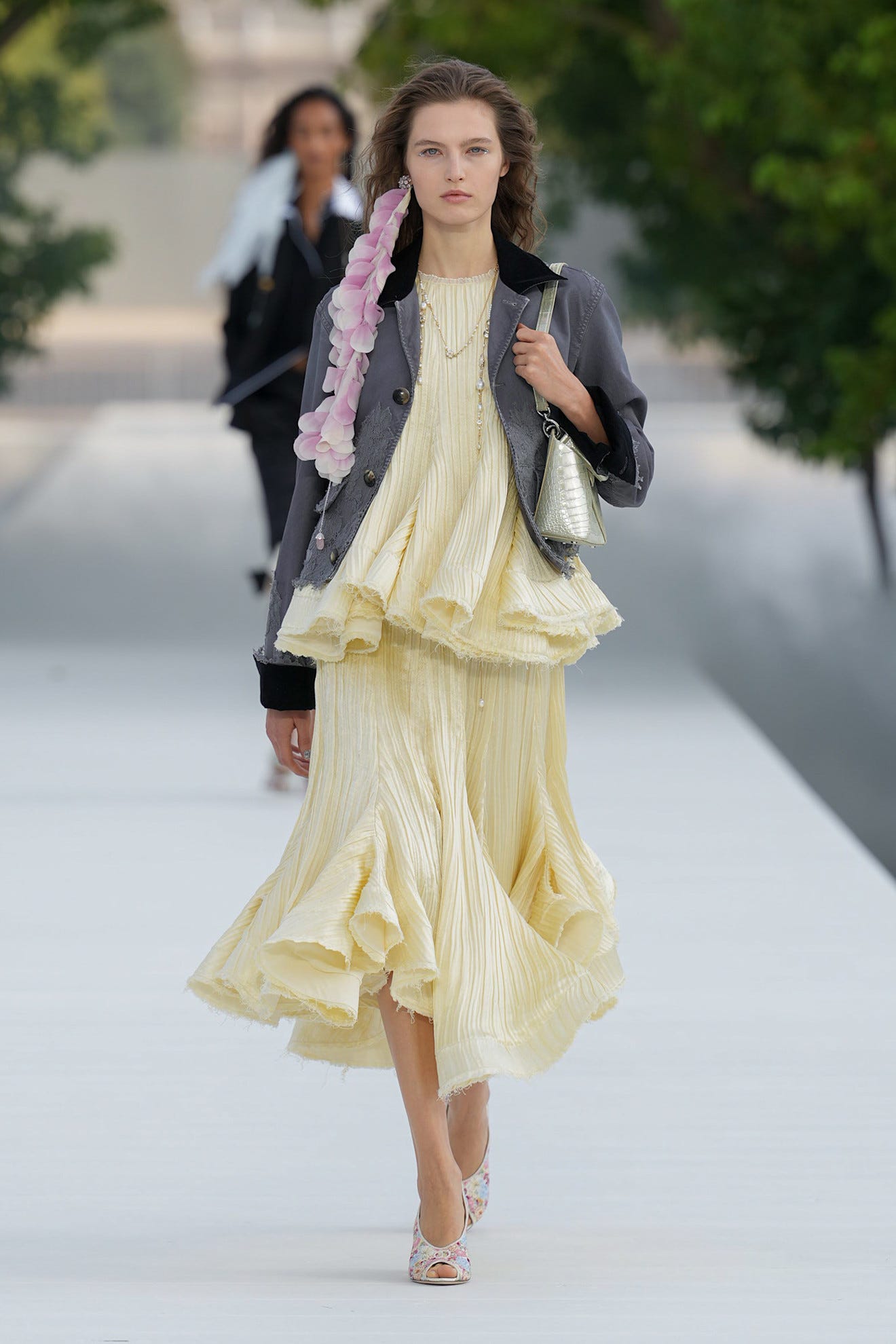 Model wearing a layered yellow dress and grey jacket, walking on a runway with lush greenery in the background.