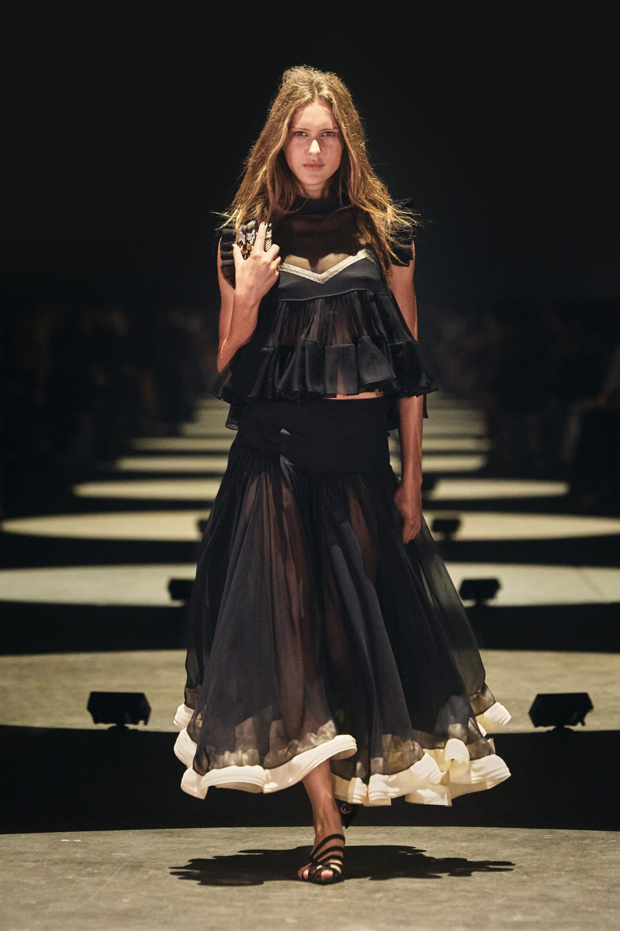 Model walking the runway in a layered black outfit with sheer detail and flowing skirt.