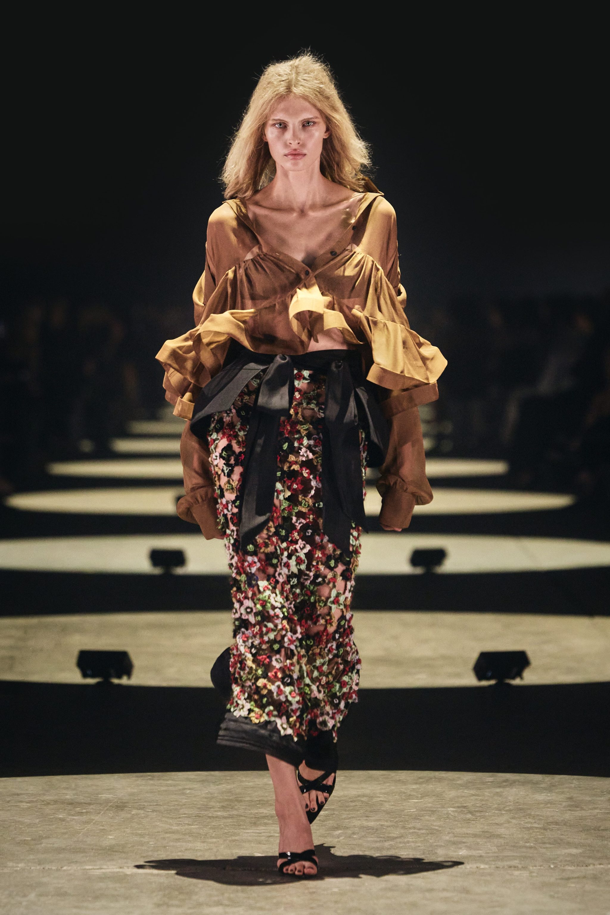 Model walking down the runway wearing a stylish outfit with a layered blouse and patterned skirt.