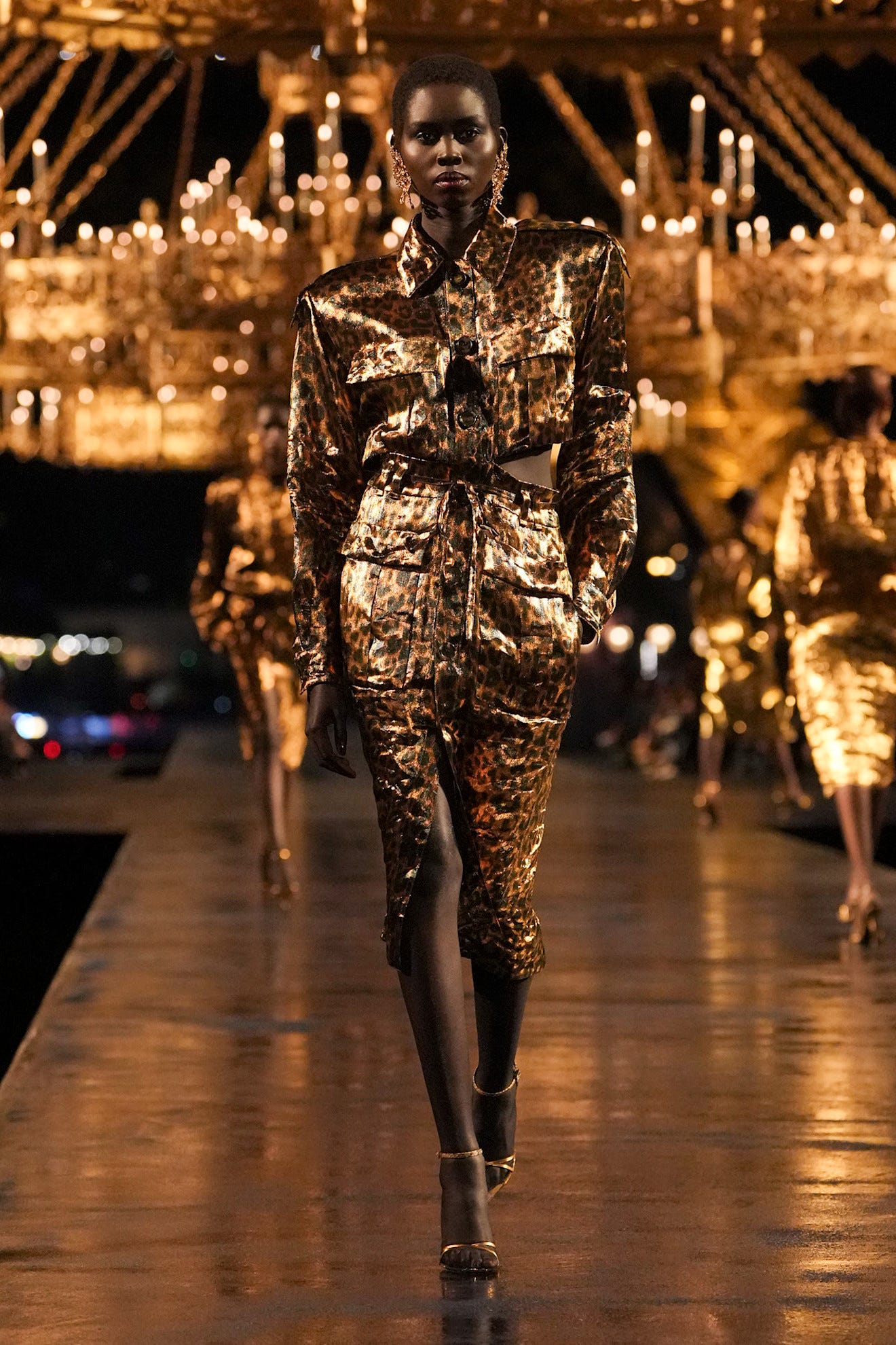Model loopt over de catwalk in een glanzende outfit met luipaardprint en kroonluchters op de achtergrond.