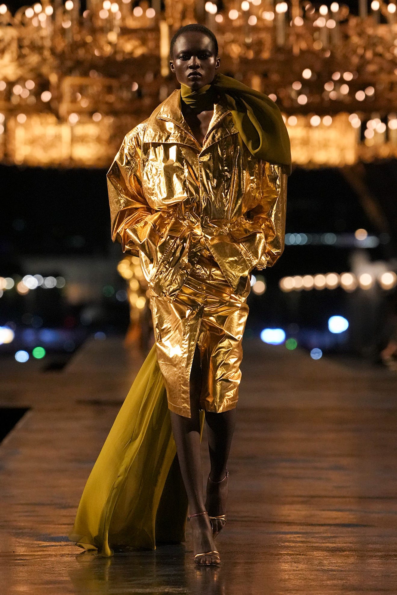 Model loopt over de catwalk in een glanzende gouden outfit met een zwierige olijfgroene sleep en hakken.