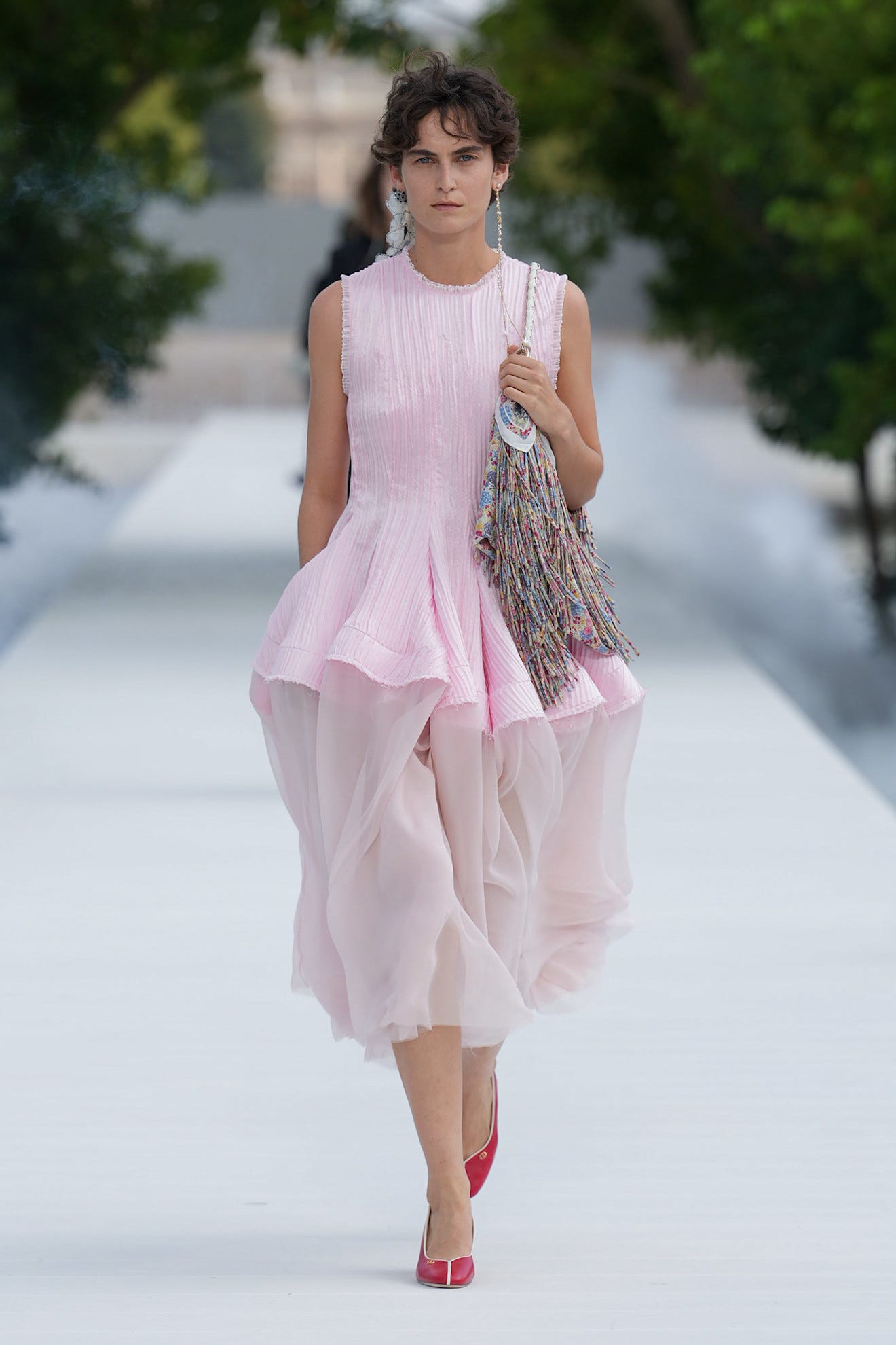 Model walking on runway in a pink dress and red shoes, holding a colorful bag, with blurred greenery in the background.