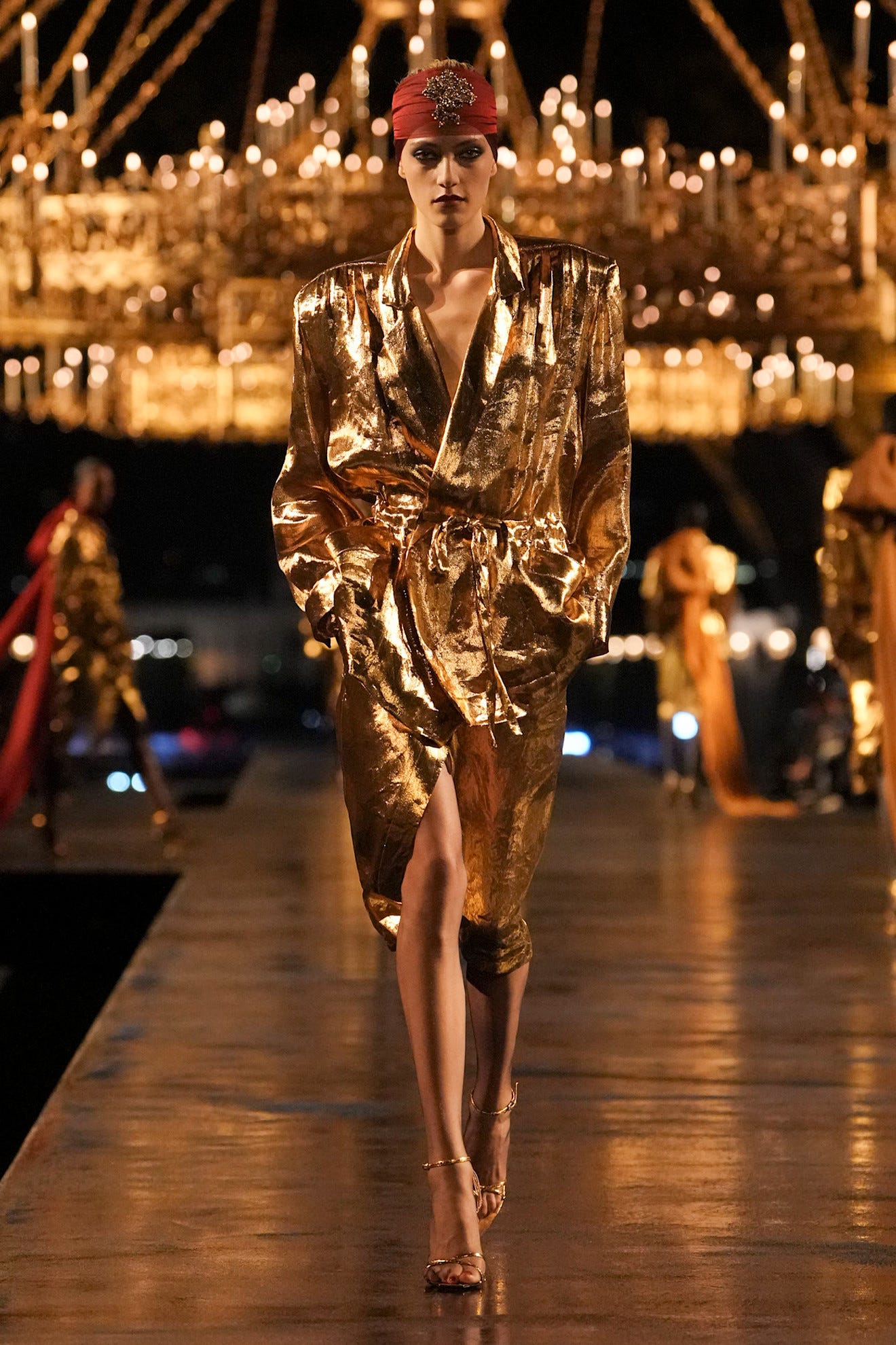 Model in een glanzende gouden mantel loopt over de catwalk, met fonkelende kroonluchters en dramatische verlichting op de achtergrond.