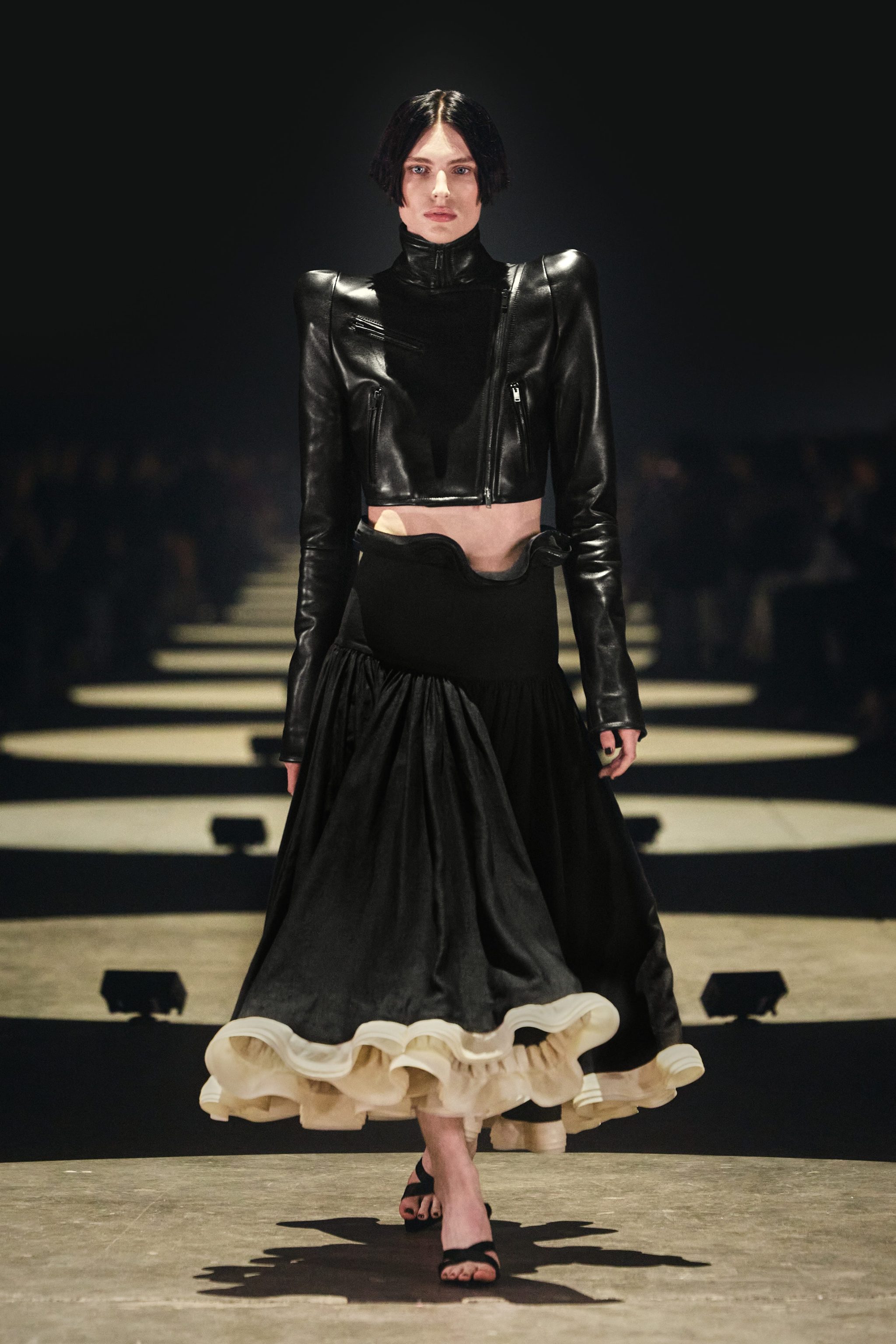 Model walking the runway in a black leather cropped jacket and long black skirt with ruffled hem, under dramatic lighting.