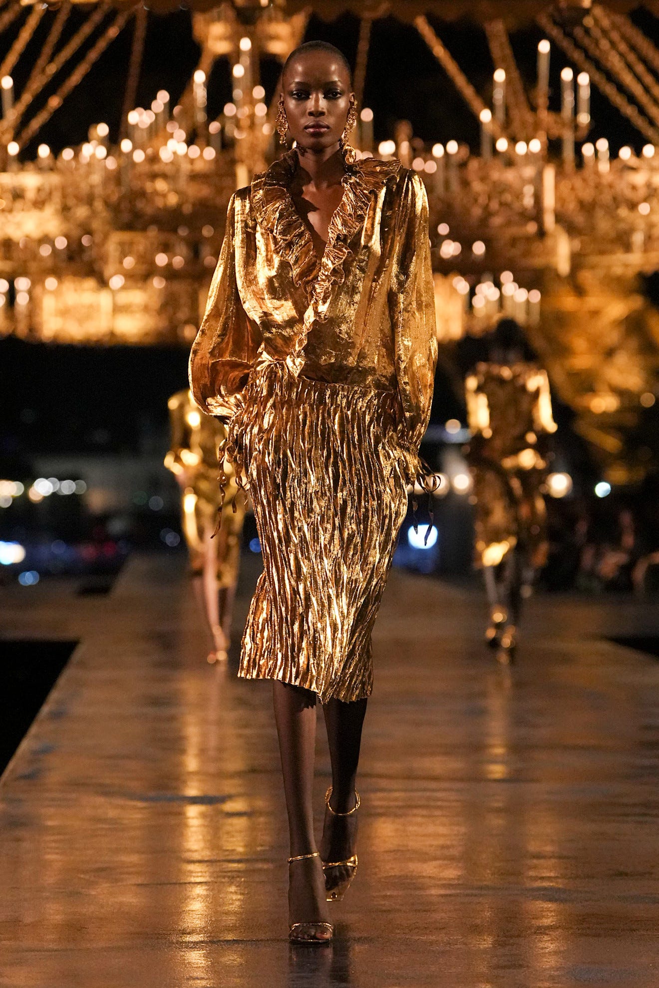Model loopt over de catwalk in een fonkelende gouden outfit met ruches en plooien, verlicht door kroonluchters.