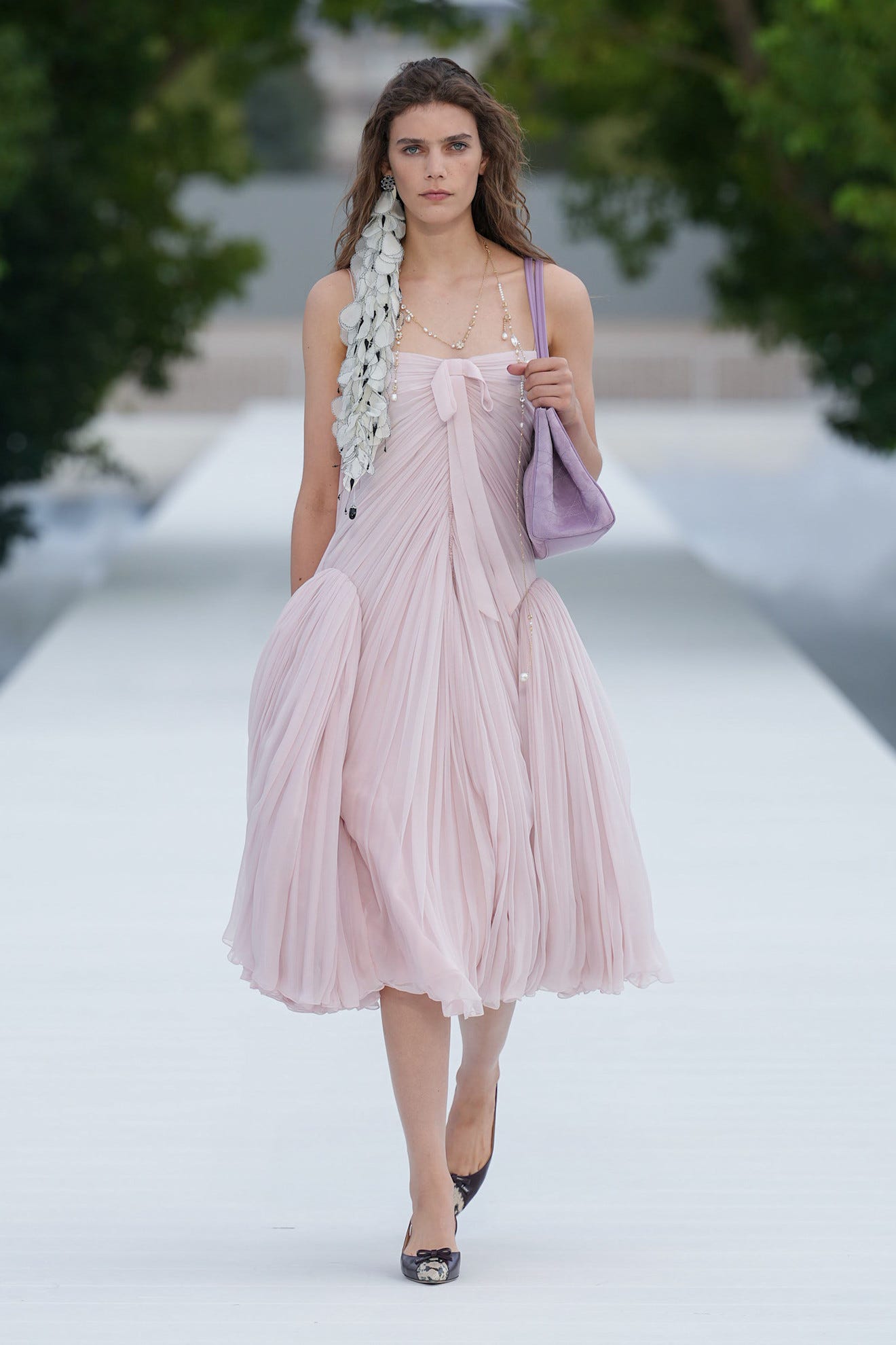 Model walking on a runway in a flowing pink dress, accessorized with a unique hairstyle and a purple handbag.