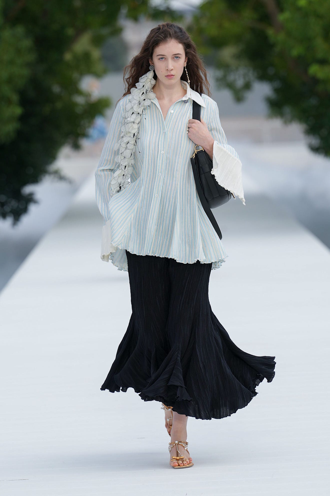 Model walking on runway in a striped shirt, black skirt, and stylish sandals, with greenery in the background.