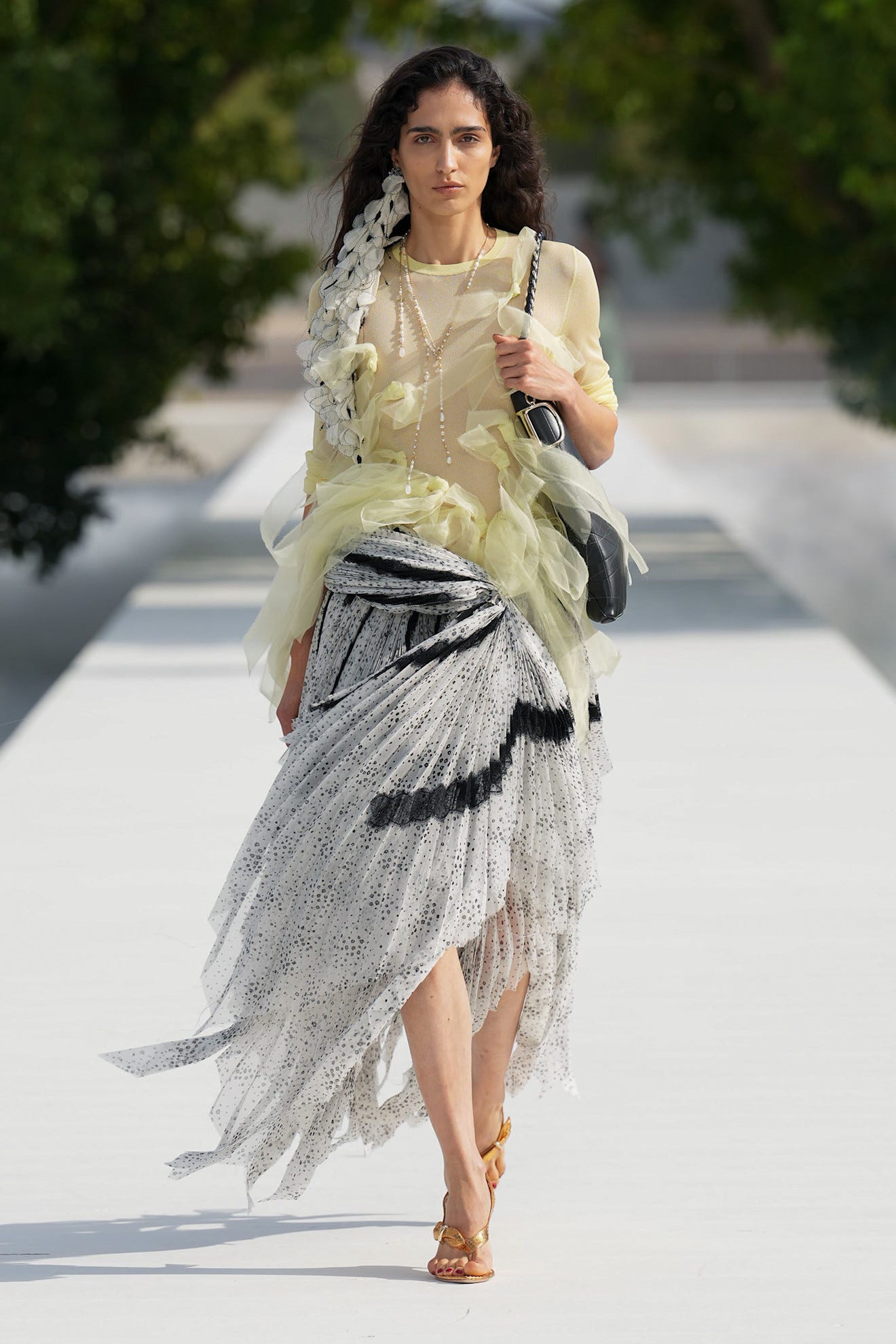 Model walking on runway wearing a sheer yellow top, black-and-white patterned skirt, and stylish accessories.
