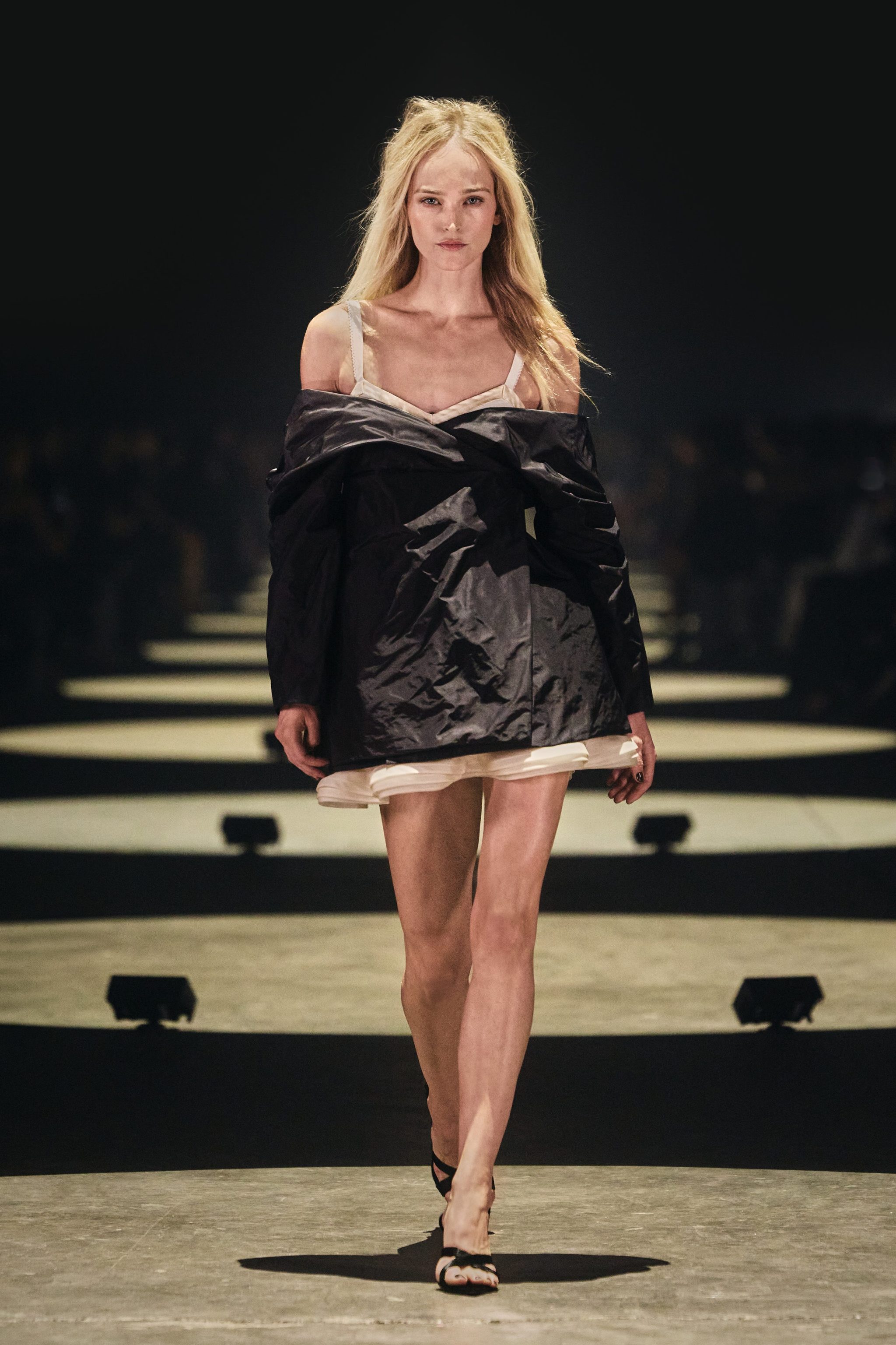 Model walking on the runway in a black oversized jacket and white dress, showcasing a fashion design.