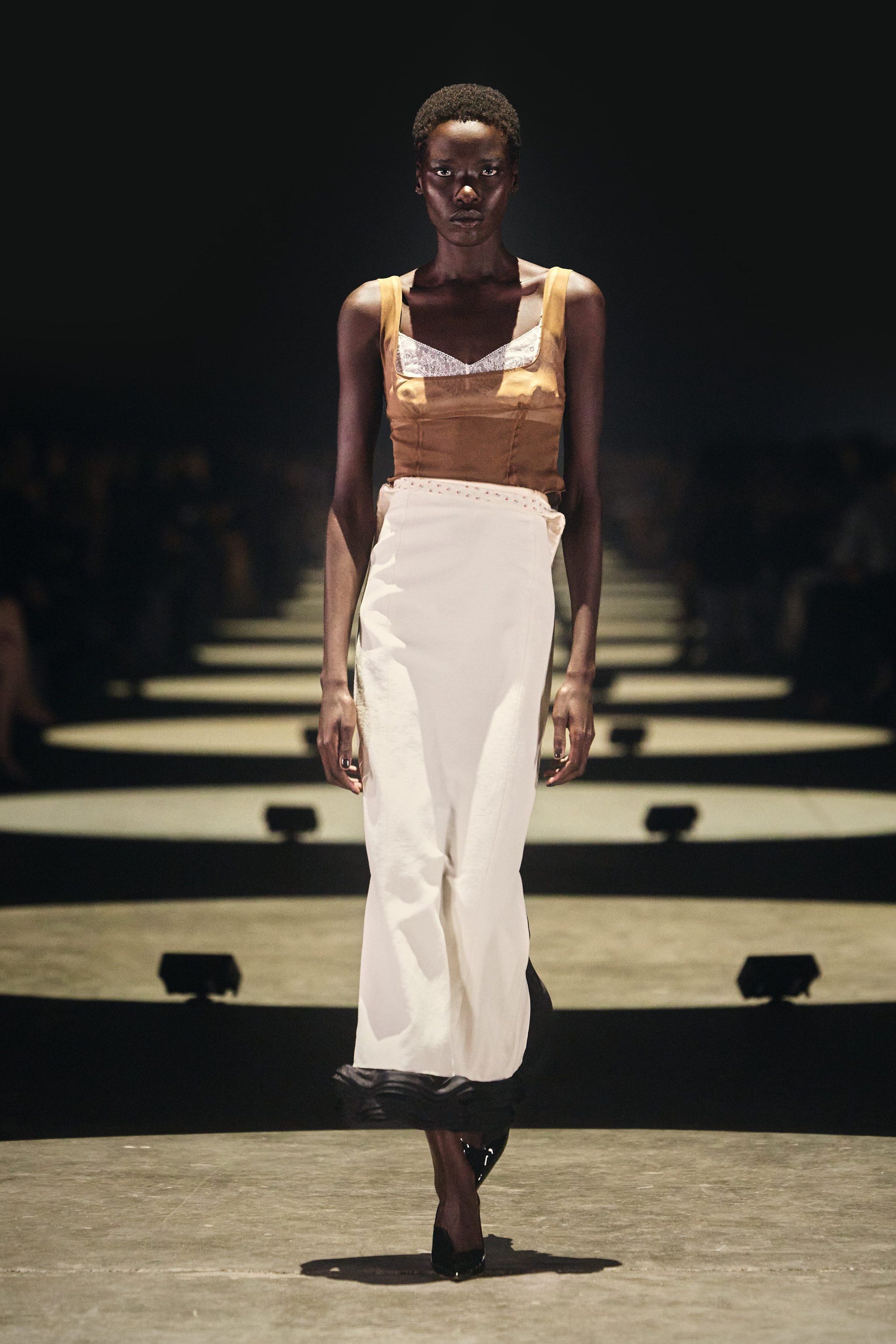Model walking down the runway wearing a brown top and white skirt, showcasing a fashion design in a dimly lit venue.
