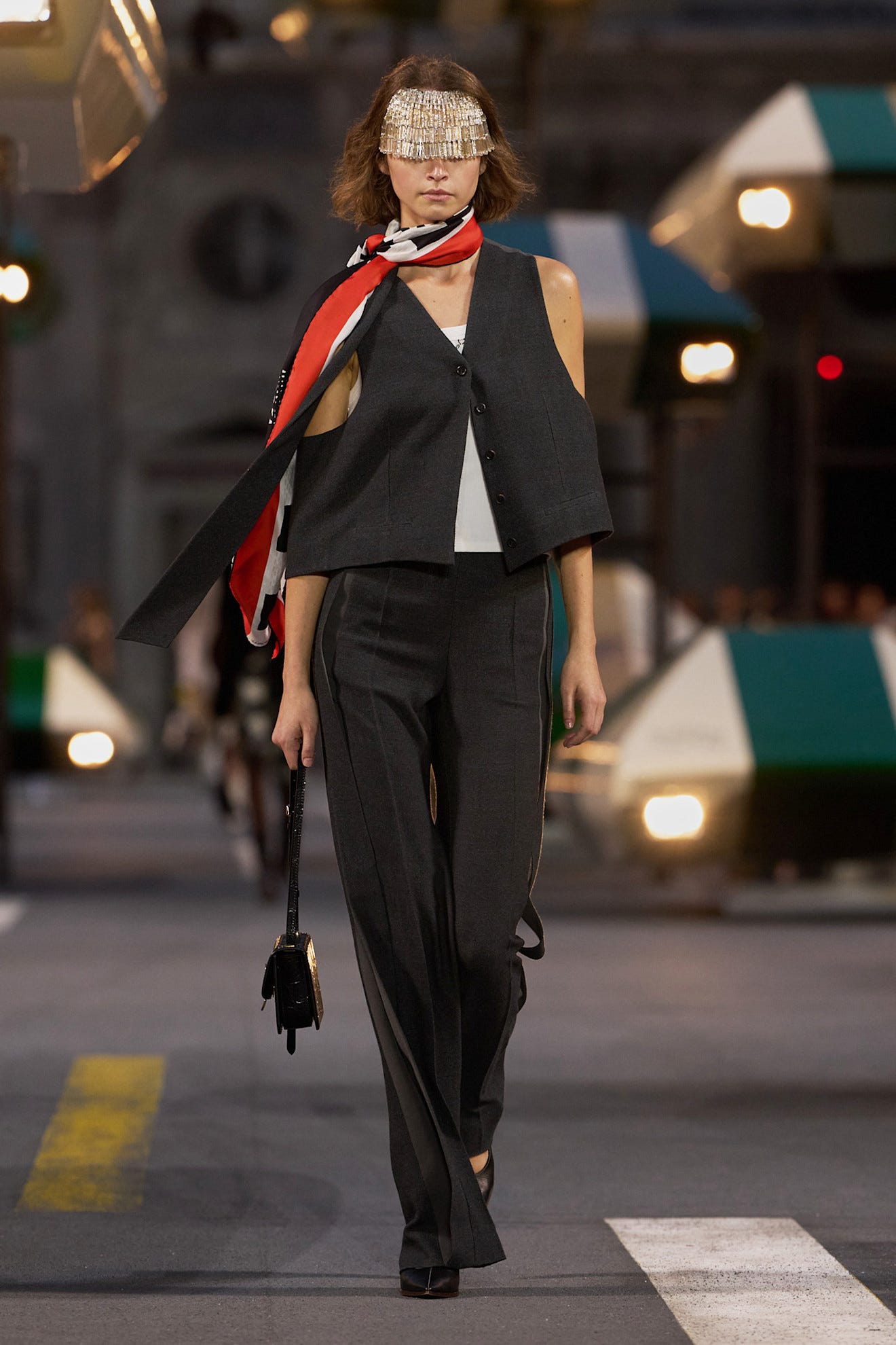 Model walking the runway in a stylish dark suit with a beaded visor and colorful scarf, holding a small black handbag.