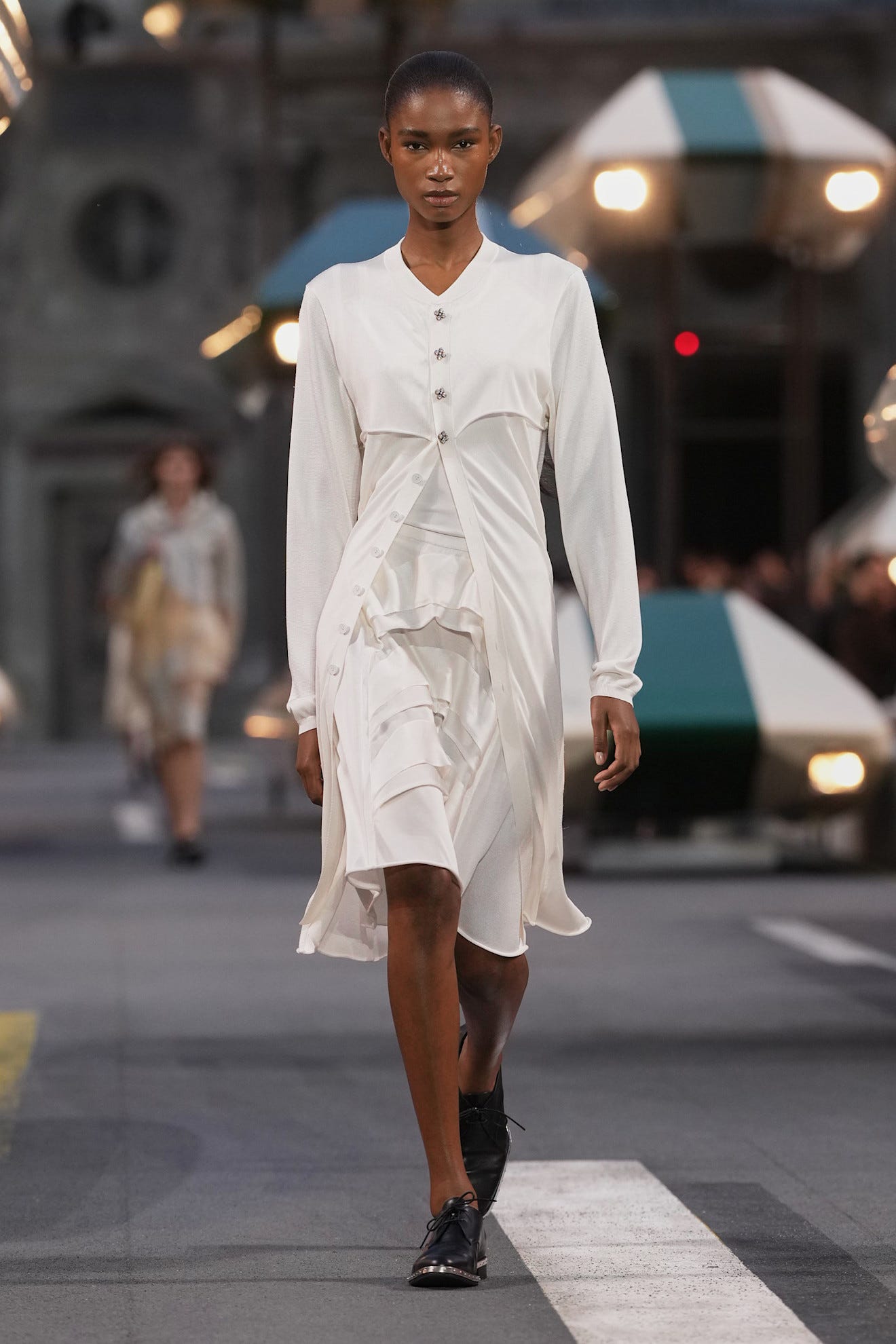 Model wearing a stylish white outfit walking down a runway with blurred background figures and contemporary set design.