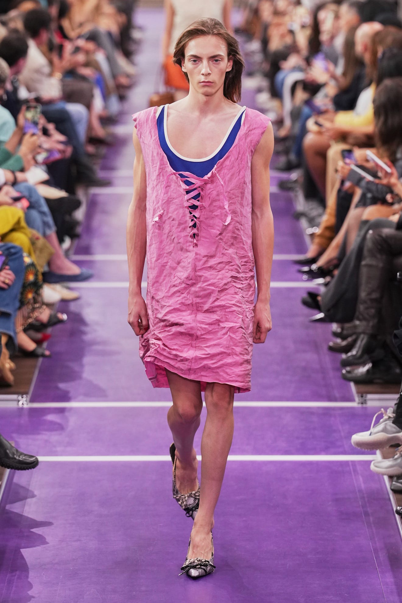 Model walking down a runway wearing a pink dress and blue undershirt, with an audience seated on either side.