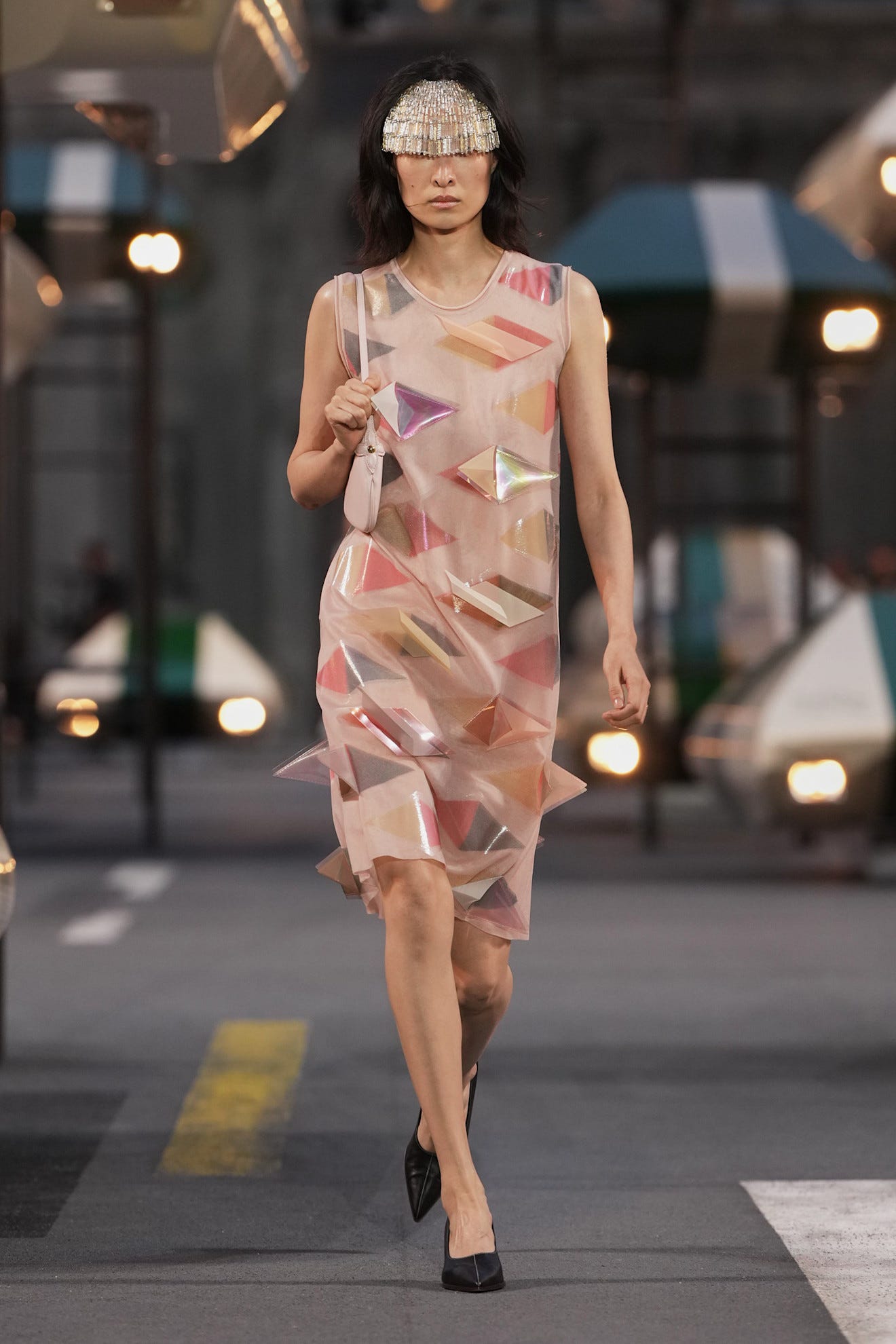 Model walking down a runway in a sheer dress adorned with colorful geometric shapes and a shimmering headpiece.