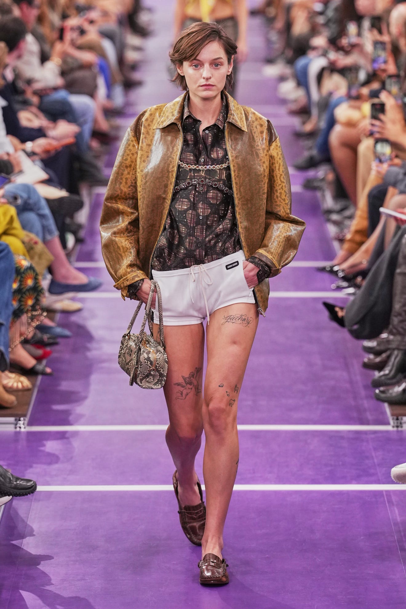Model walking the runway in a stylish outfit featuring a brown jacket and white shorts during a fashion show.