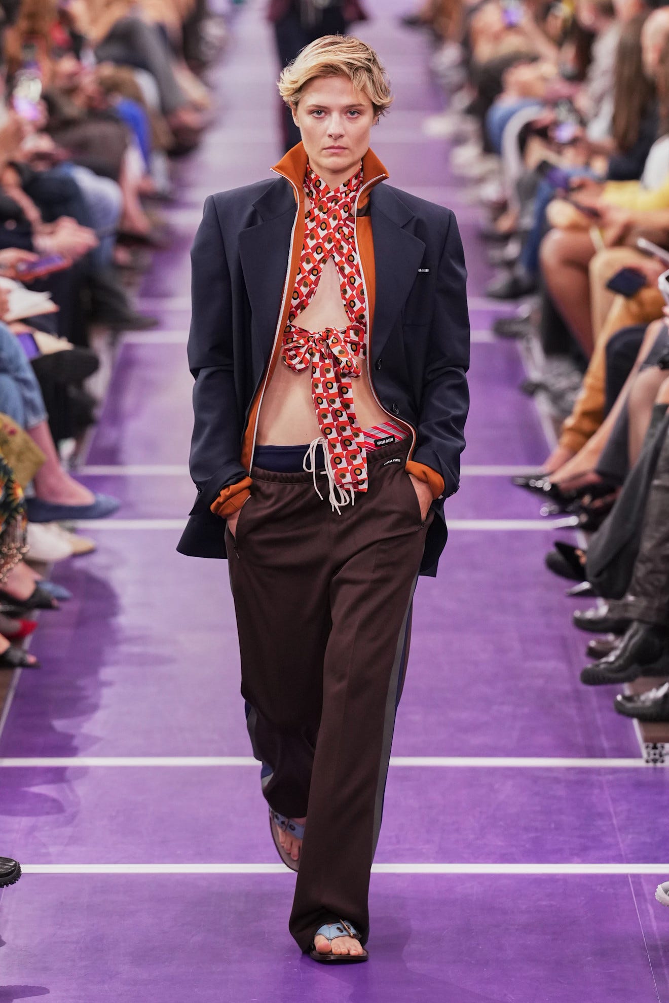 Model walking the runway in a bold, layered outfit featuring a patterned scarf and casual pants at a fashion show.