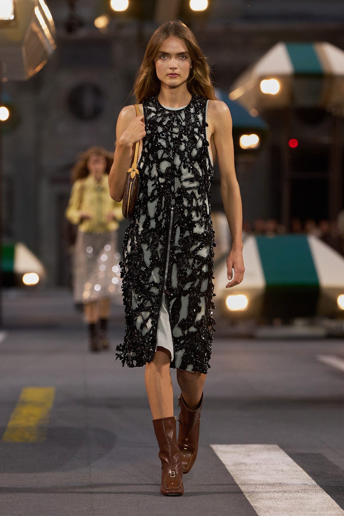 Model walking in a stylish black and white dress with intricate detailing, paired with brown boots on a runway.