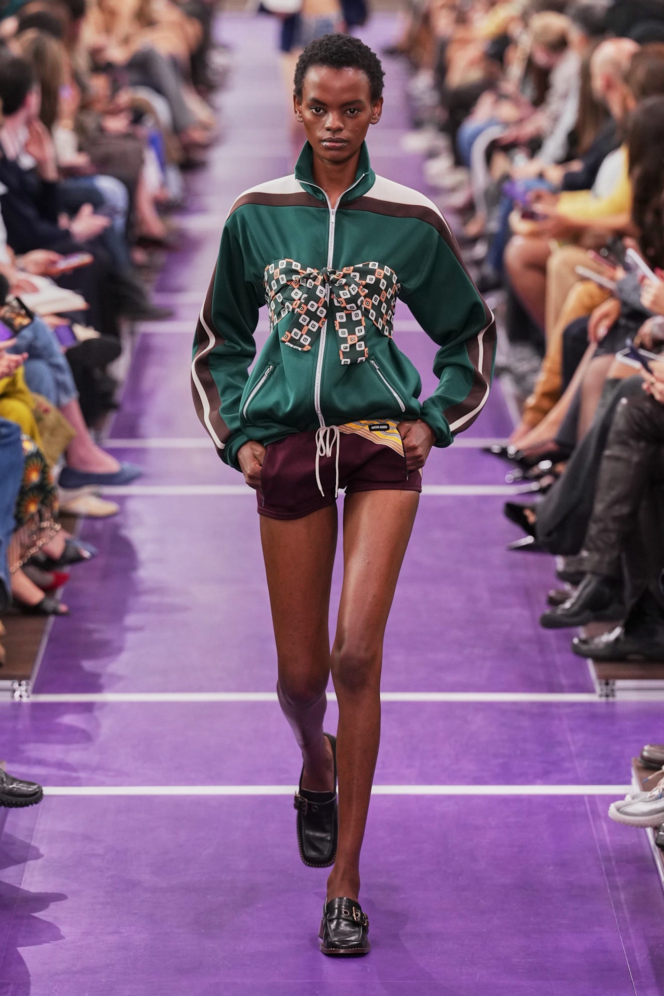 Model walking the runway in a green jacket with decorative bow and maroon shorts during a fashion show.