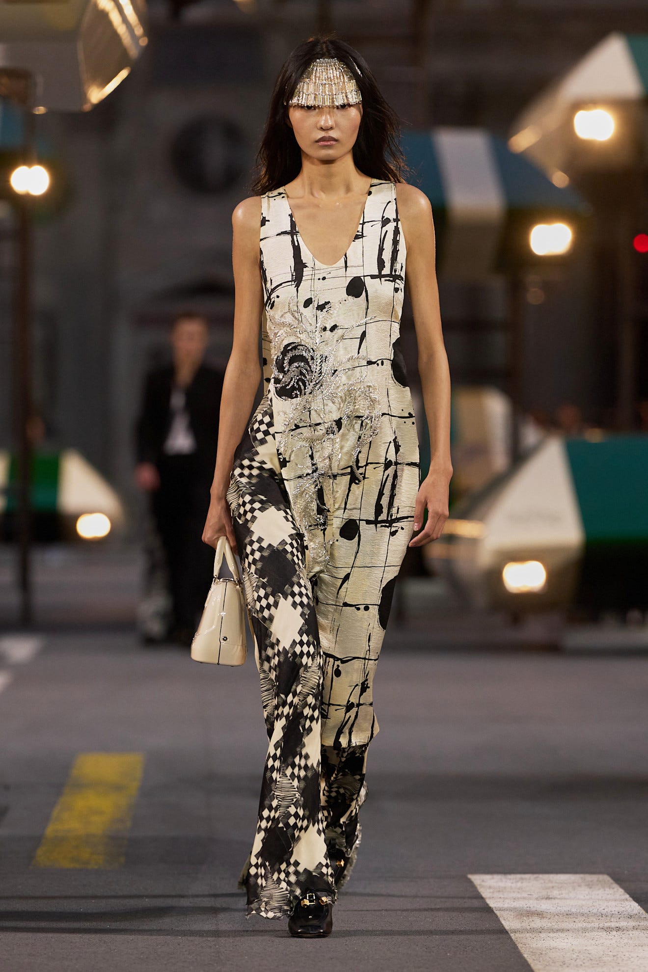 Model wearing a stylish abstract-pattern jumpsuit and a unique headpiece, walking on a runway with illuminated backdrop.