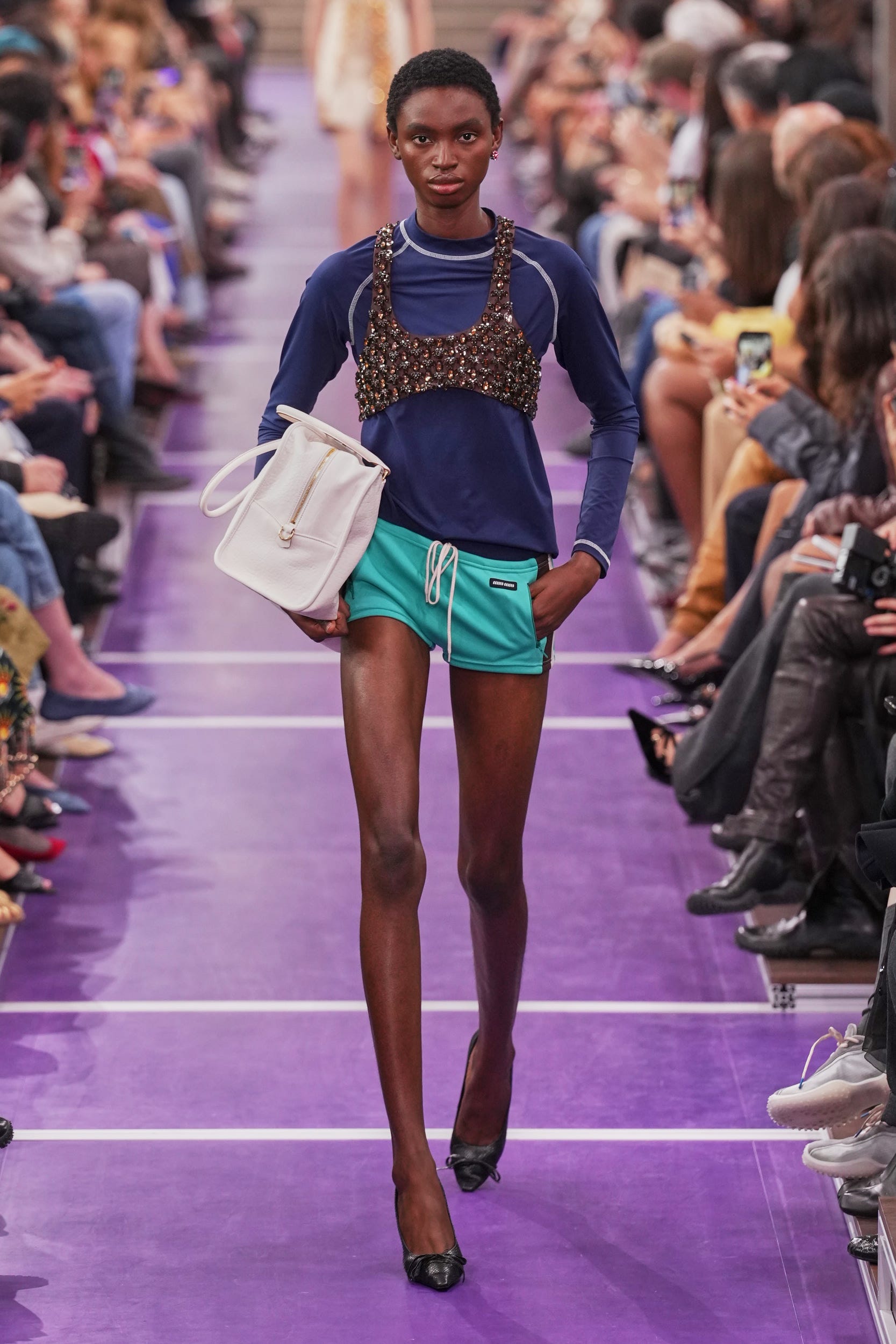Model walking down the runway in a blue top, embellished crop, green shorts, and carrying a white handbag.