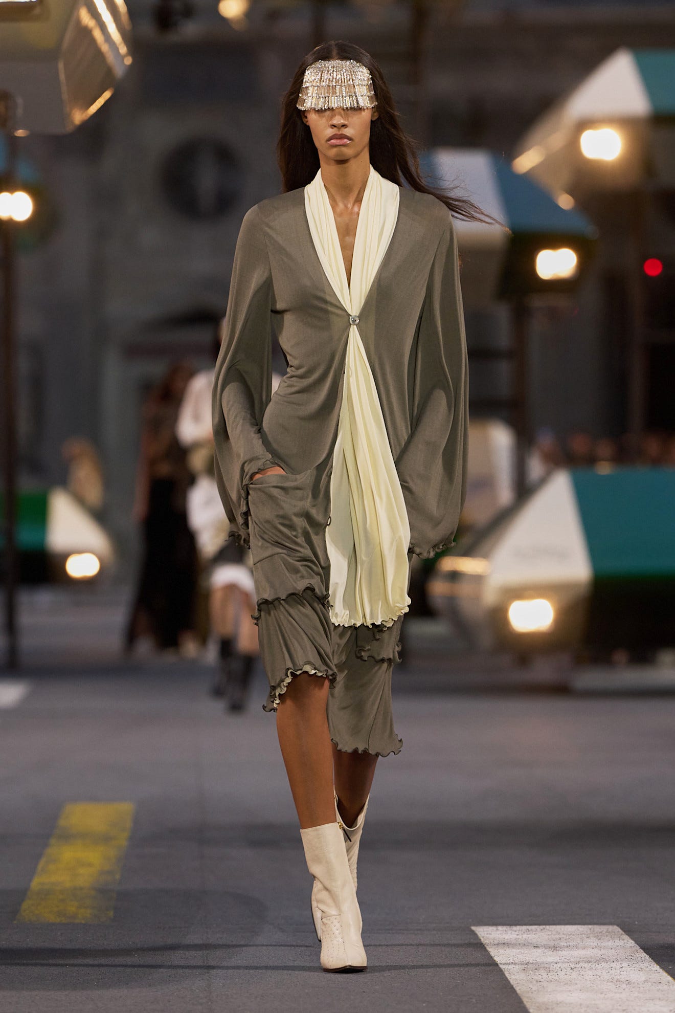 A model walks the runway wearing a grey outfit with a white scarf and a beaded visor in an outdoor fashion show setting.